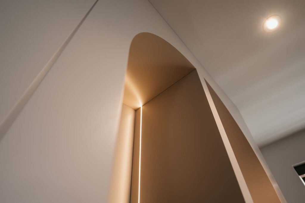 Minimalistisch architectonisch element met een zacht verlichte, gebogen boog en vloeiende, schuine wanden. Warm licht accentueert de boog en zorgt voor dramatische schaduwen en een serene, moderne sfeer in een interieur met neutrale tinten.