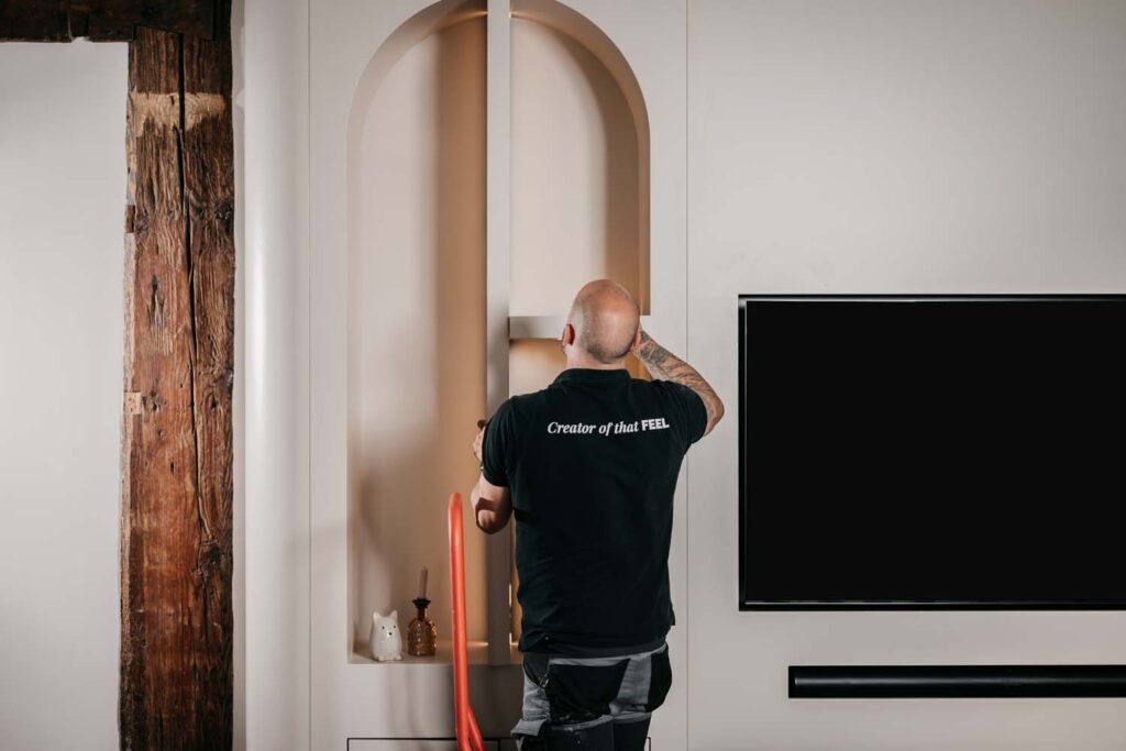 Een persoon met tatoeages, gekleed in een zwart T-shirt met de tekst ‘Creator of that FEEL’, is bezig met een moderne beige wandkast naast een flatscreen-tv en een zichtbare houten balk.