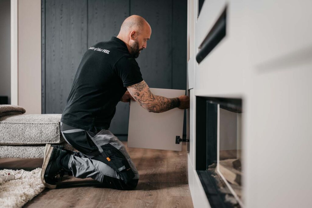 Een persoon met tatoeages zit op de grond terwijl hij een kastdeur monteert of repareert in een modern interieur.